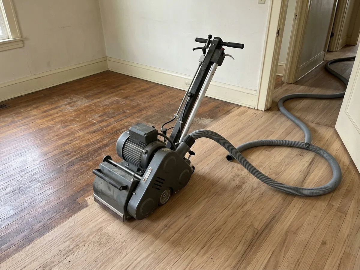 Belt floor sander with a thick hose running from its dust port across a half-sanded oak floor