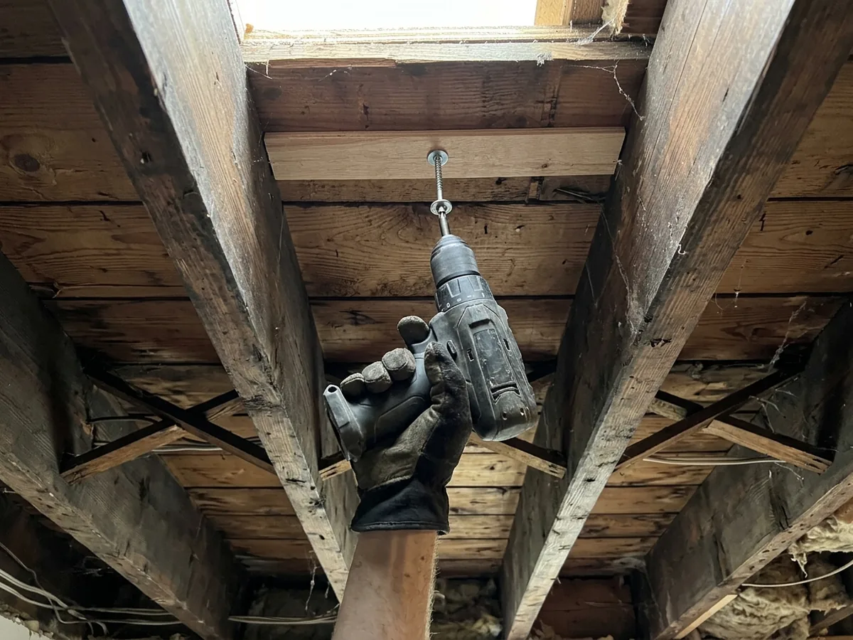Hand driving a screw up through a squeaky subfloor into the floor above, seen from the basement between wood joists